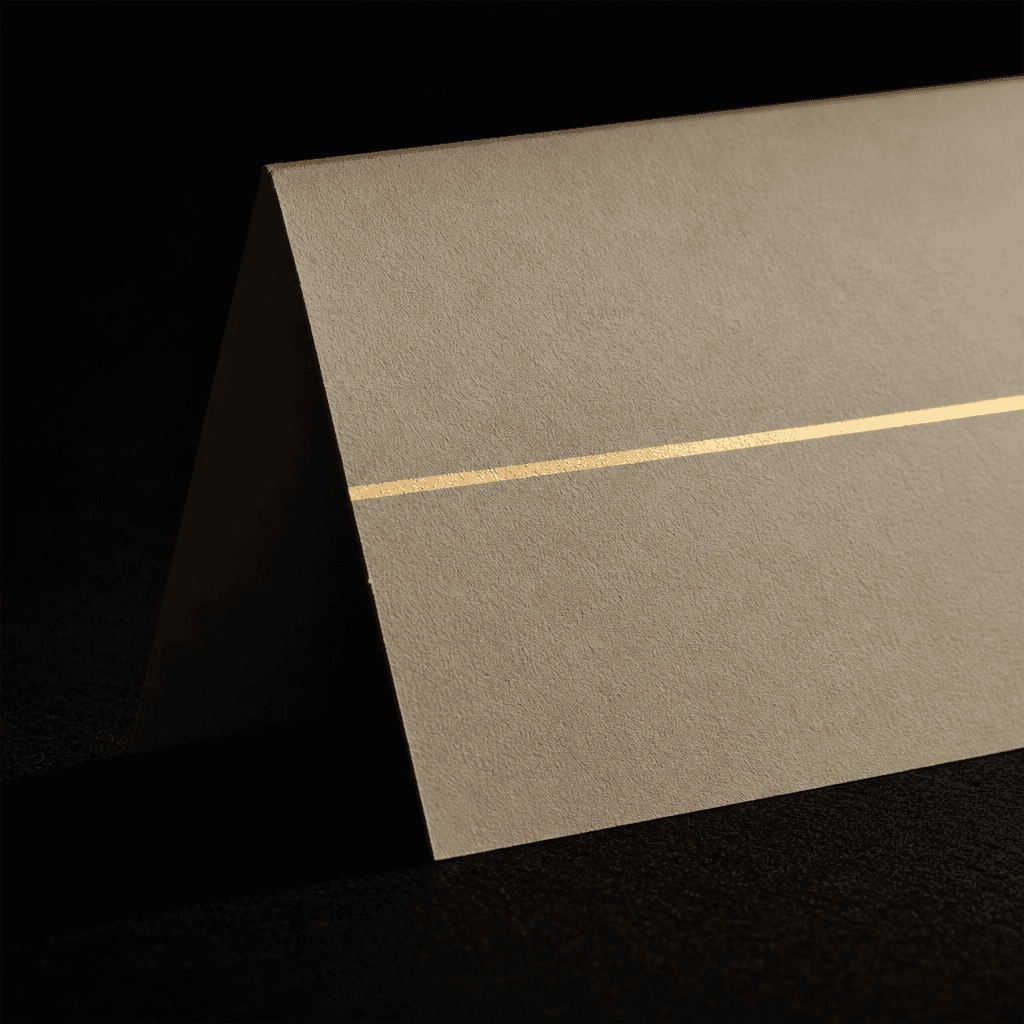 A single thin brass line of light falling across a folded cream card on a black surface